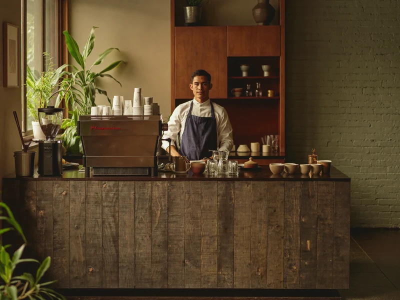 Illustrierter Innenraum eines kleinen Kaffeehauses mit Barista am Holztresen im Morgenlicht
